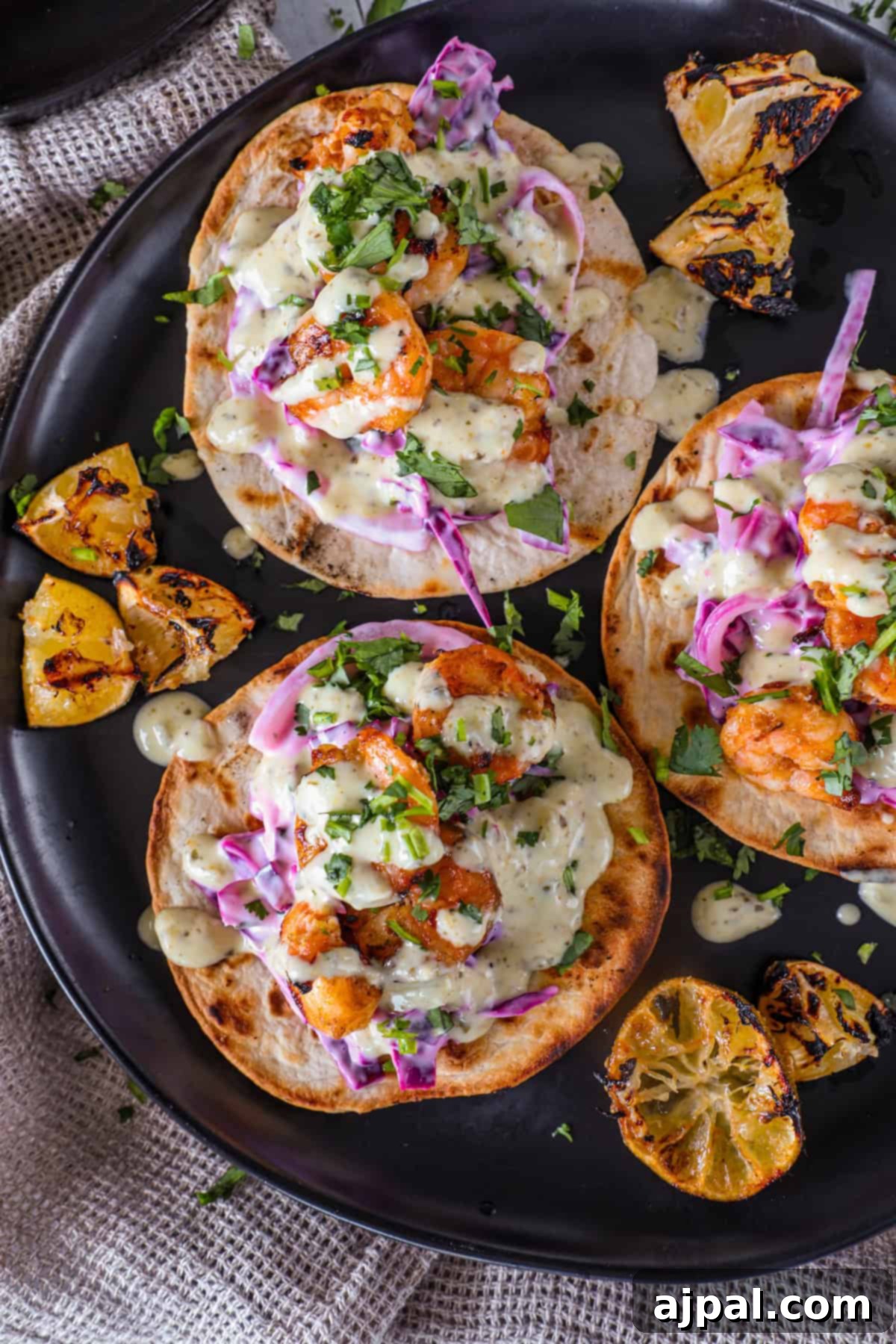 Coastal Shrimp Tacos 2 Three grilled shrimp tacos garnished with vibrant purple cabbage slaw, fresh cilantro, and a creamy sauce, served alongside grilled lemon wedges on a sleek black plate.