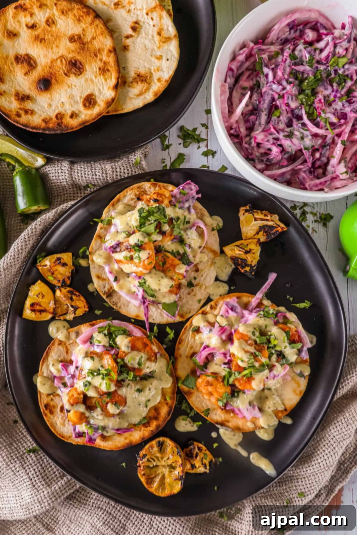 Coastal Shrimp Tacos 3 Three artfully arranged shrimp tacos, featuring cilantro lime crema, on a black plate. The scene is completed with charred lemon halves, extra tortillas, and a bowl of fresh coleslaw in the background.