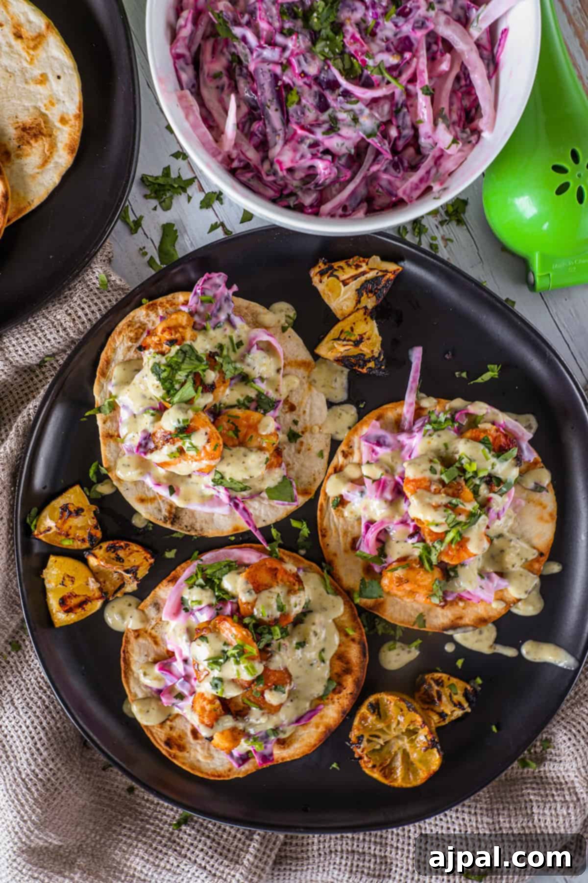 Coastal Shrimp Tacos 6 An overhead shot showcasing three perfectly plated shrimp tacos, topped with creamy sauce, a generous helping of purple cabbage slaw, and fresh herbs. Charred lime wedges accompany the tacos, with a green juicer visible in the soft background.