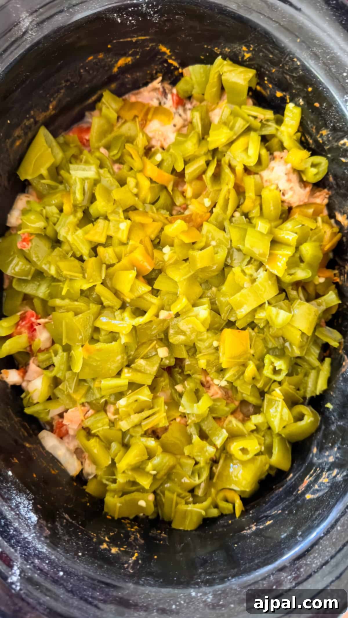 A slow cooker filled with floured pork, diced onions, garlic, and vibrant roasted green chiles, illustrating the early stages of preparing a Colorado pork green chile.