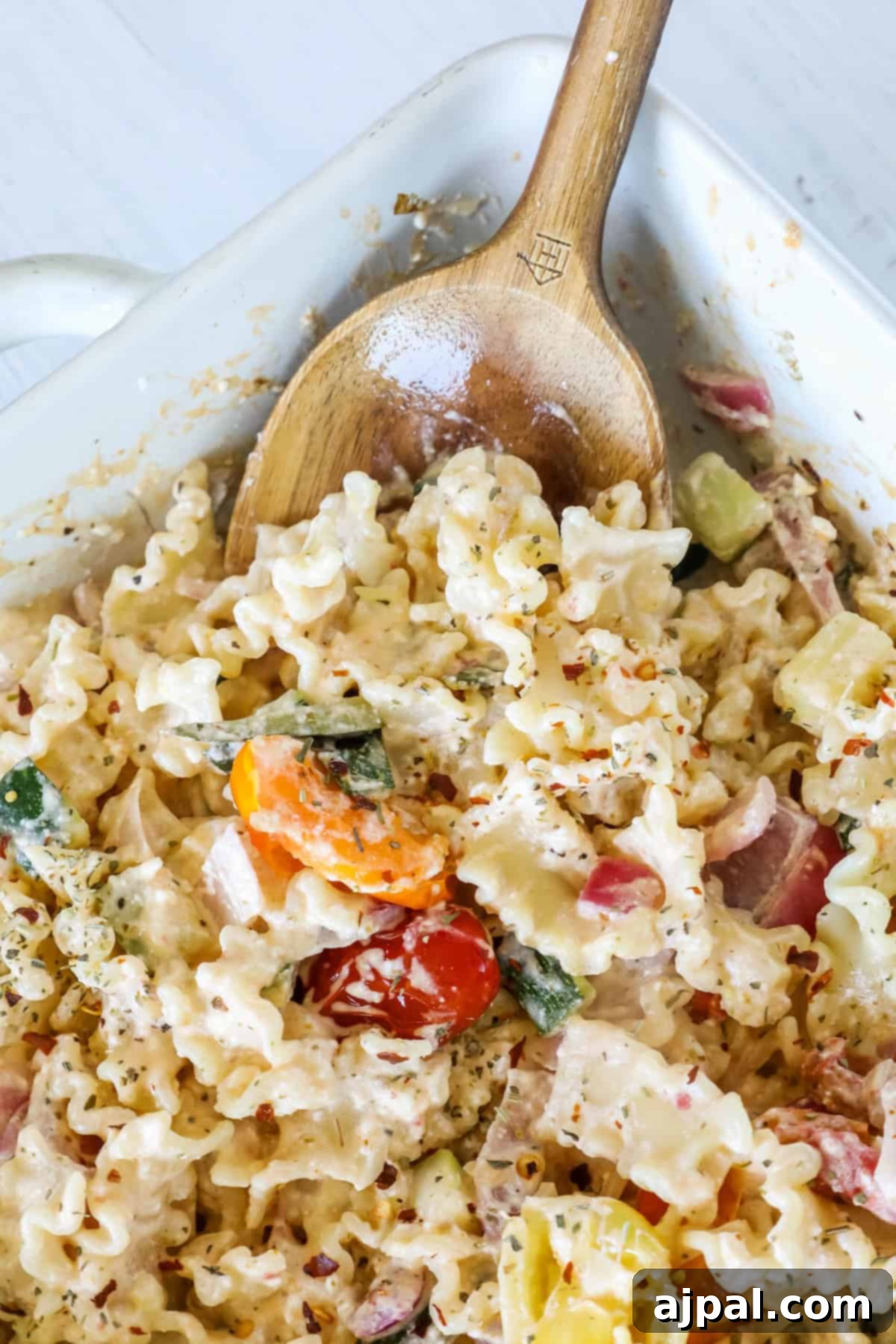 Baked Feta Pasta Bliss 12 Close-up of a pasta dish with a creamy sauce, mixed with vegetables such as tomatoes, zucchini, and bell peppers, served in a white casserole dish with a wooden spoon.