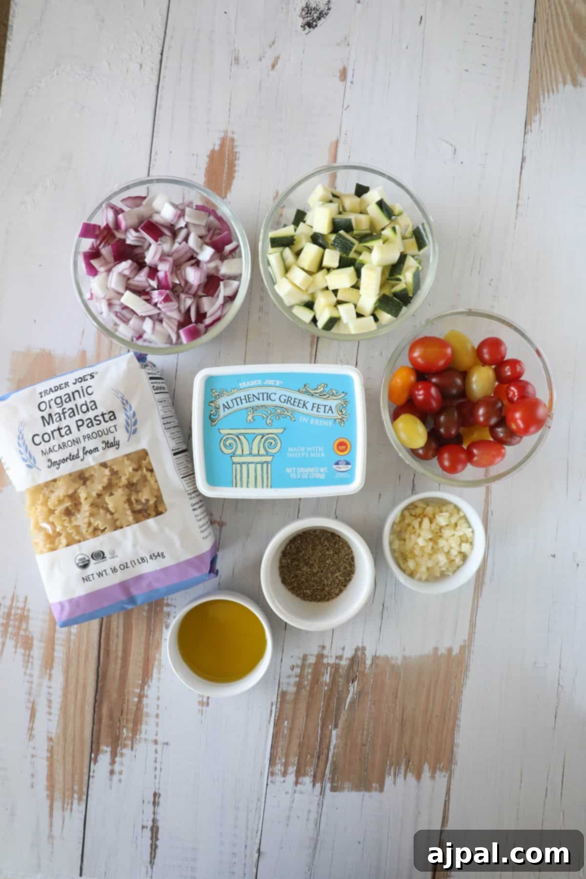 Baked Feta Pasta Bliss 5 Ingredients for a meal arranged on a wooden surface, including chopped red onion, chopped zucchini, cherry tomatoes, Greek feta cheese, Mafalda pasta, olive oil, garlic, and a spice blend.