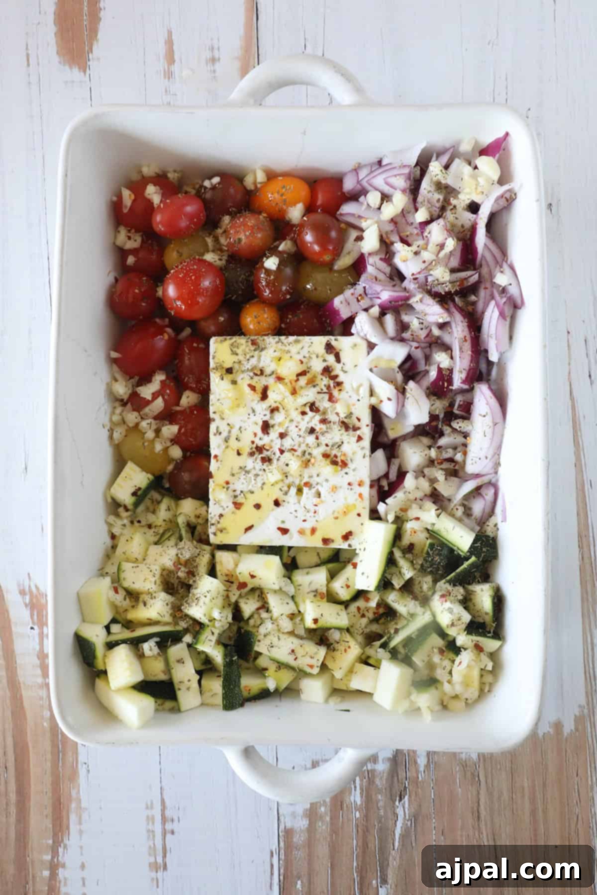 Baked Feta Pasta Bliss 6 A baking dish filled with chopped tomatoes, red onions, zucchini, garlic, and a block of feta cheese, all seasoned with spices.