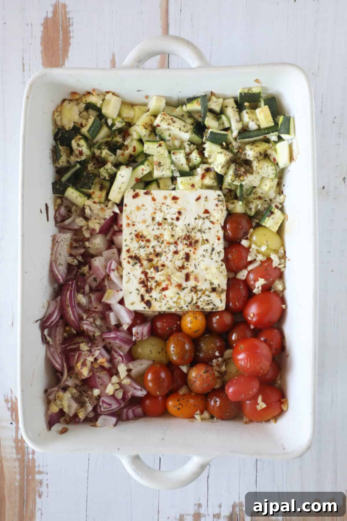 Baked Feta Pasta Bliss 7 A baking dish containing chopped zucchini, tomatoes, red onions, garlic, and a block of feta cheese, all seasoned with herbs and ready to be cooked.