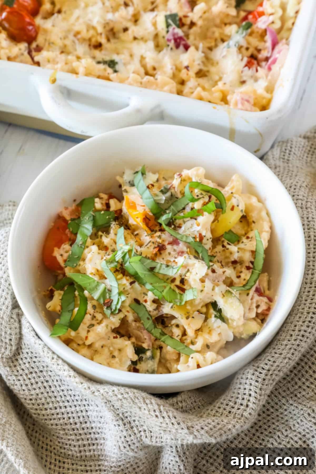 Baked Feta Pasta Bliss 9 A white bowl of baked pasta mixed with vegetables, topped with fresh basil, sits on a textured cloth near a baking dish containing more pasta.
