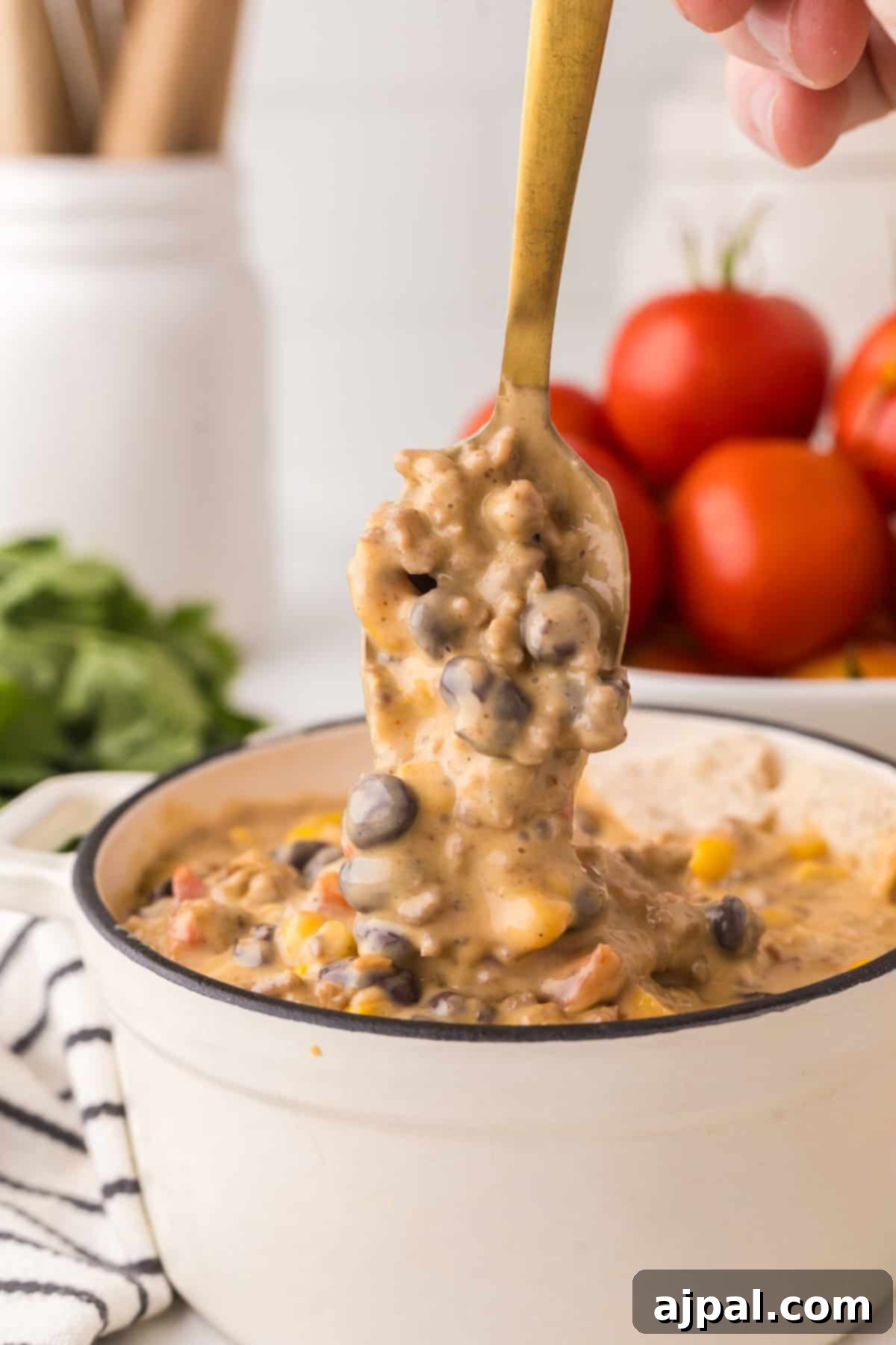A spoon is being used to lift a portion of creamy soup with black beans, corn, diced tomatoes, and a hint of queso from a pot. In the background, there is a bunch of fresh tomatoes and greens.