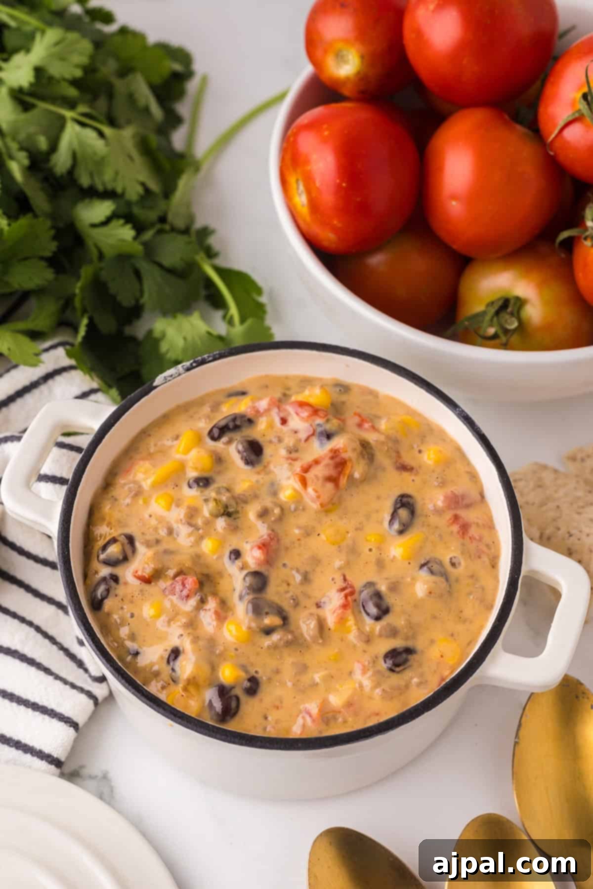 A white pot filled with a creamy Cowboy Queso soup that includes corn, black beans, diced tomatoes, and other vegetables. Fresh tomatoes, cilantro, and a striped cloth are nearby on a marble countertop.