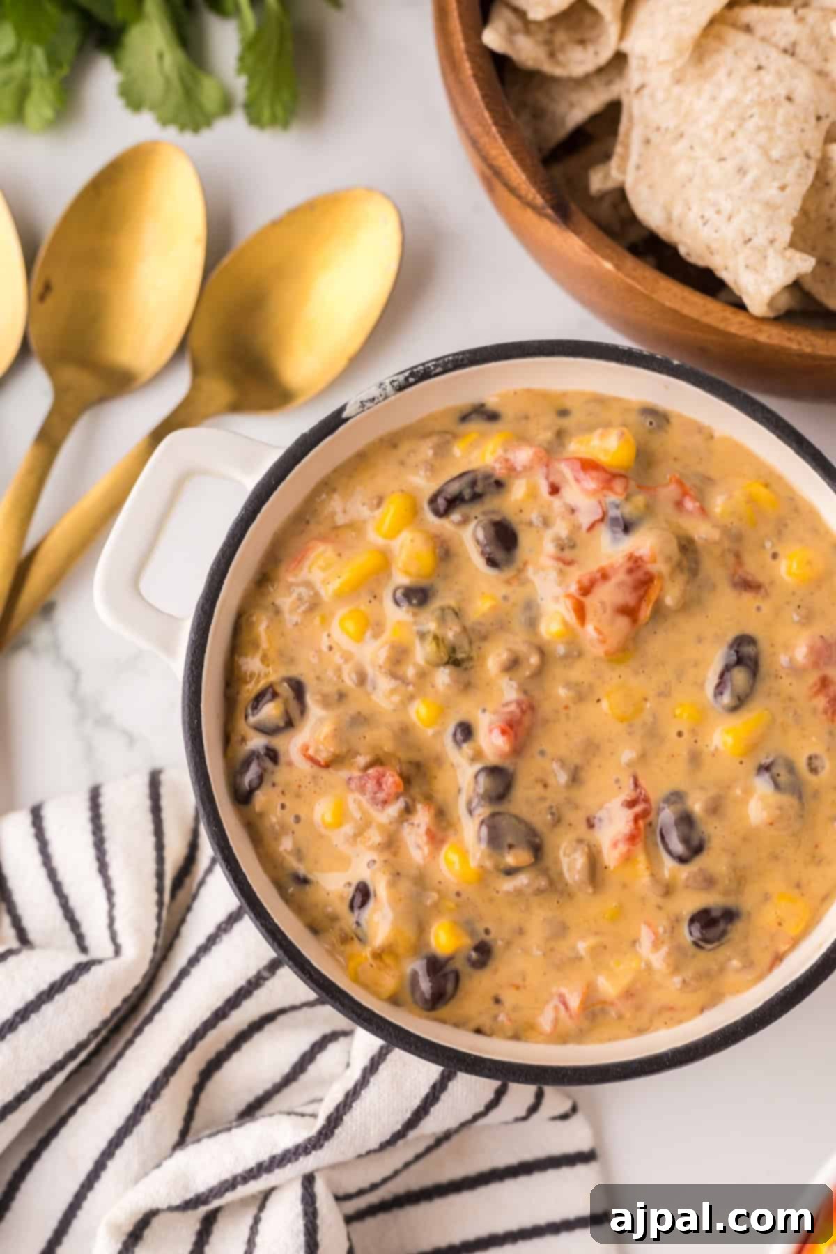 A bowl filled with creamy taco soup containing corn, black beans, diced tomatoes, and other ingredients sits next to a small dish of Cowboy Queso. Gold-colored spoons and tortilla chips in a wooden bowl are placed nearby on a marble countertop.