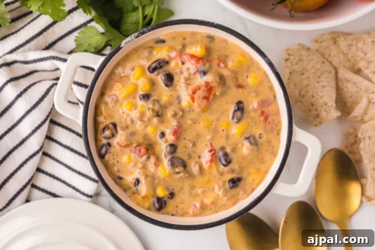 A pot of creamy soup featuring black beans, corn, and chopped tomatoes surrounded by a striped cloth, cilantro, tortilla chips, and gold-colored spoons gives way to a hint of Cowboy Queso for an unexpected kick.