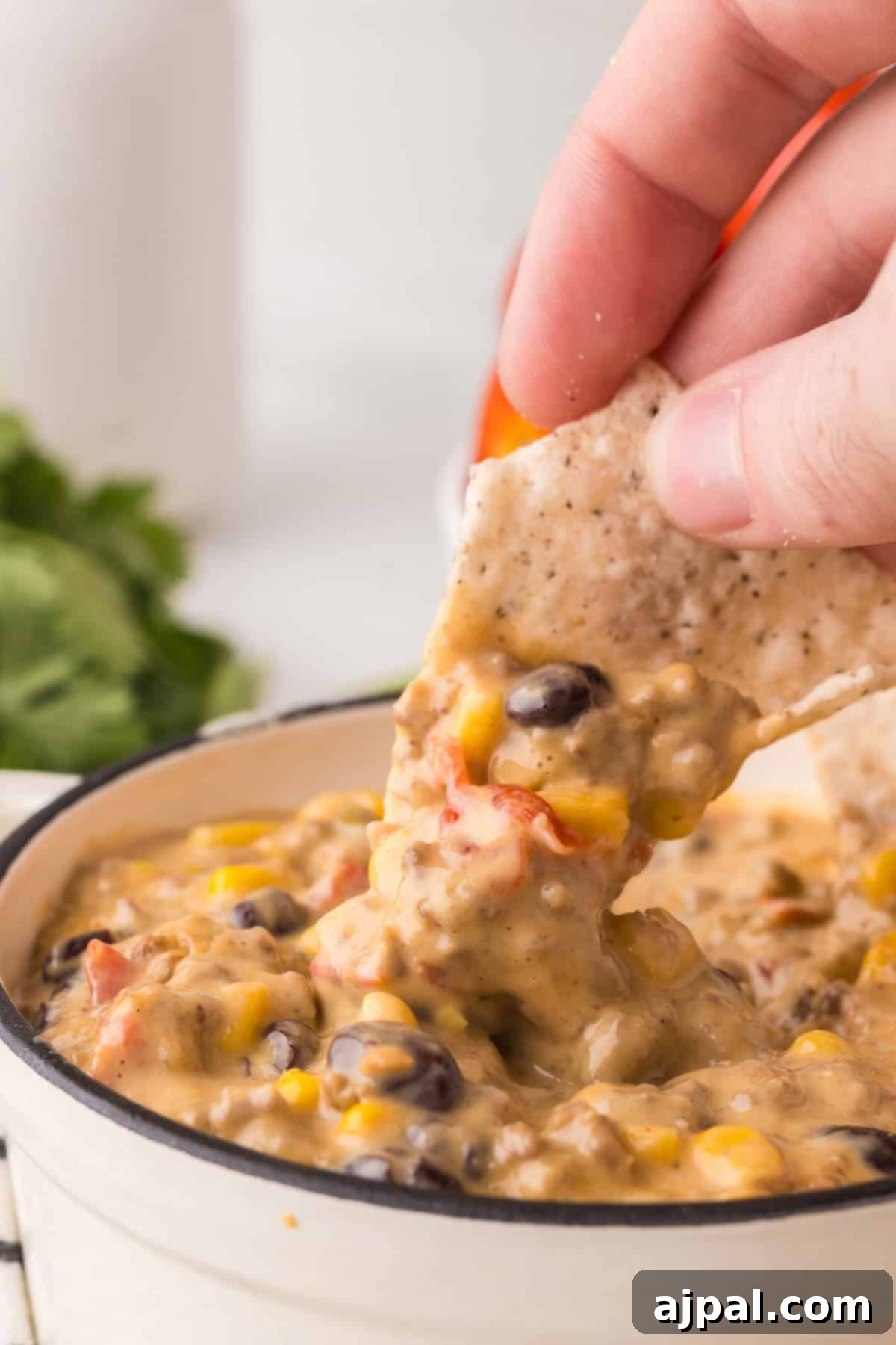 A hand dips a tortilla chip into a bowl of Cowboy Queso, a spicy cheese dip filled with black beans, corn, and red bell peppers.