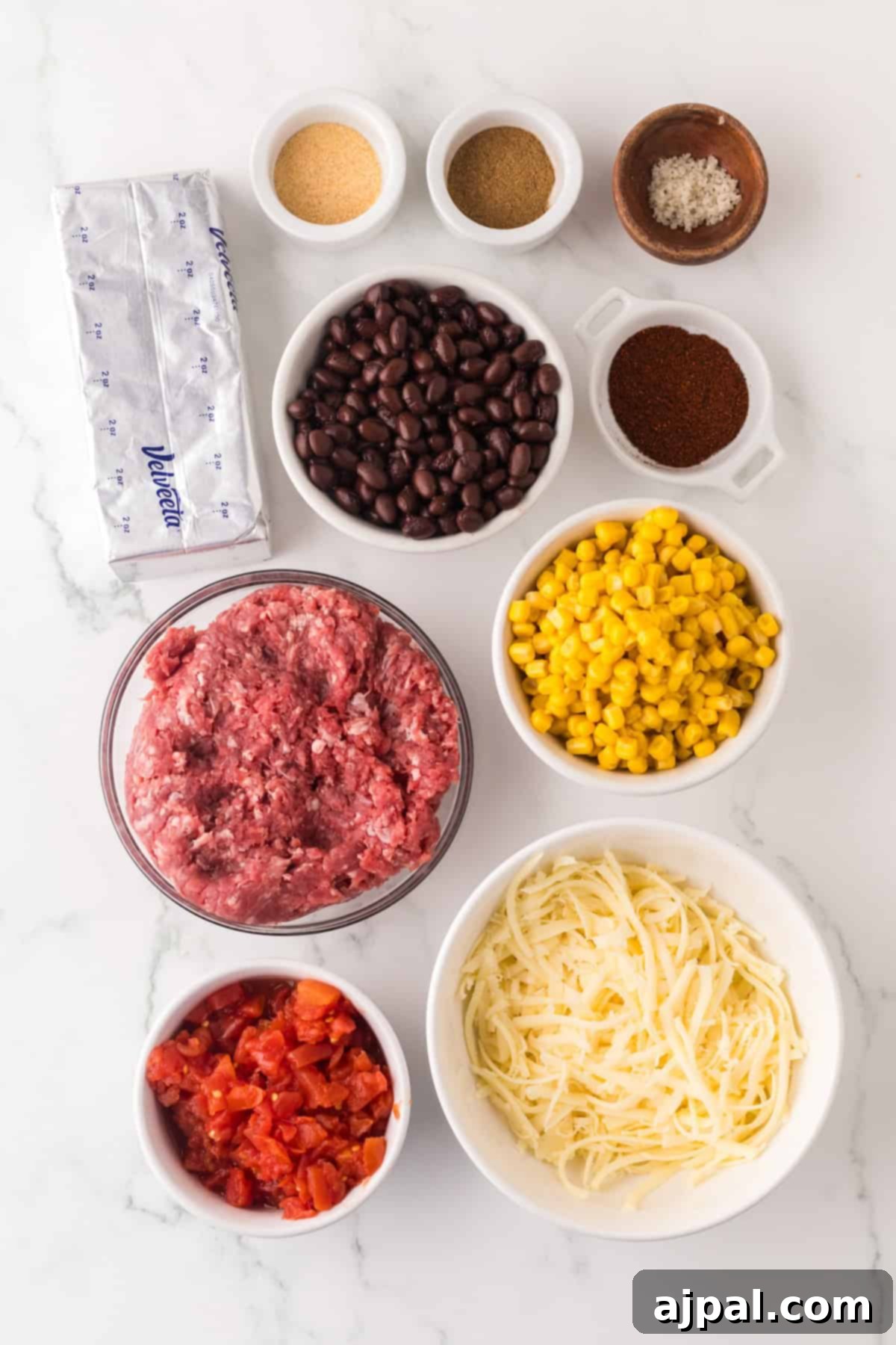 Ingredients arranged on a white surface: a block of cream cheese, ground meat, black beans, corn, shredded cheese, diced tomatoes, and various spices in small bowls—everything you need for a mouth-watering Cowboy Queso.