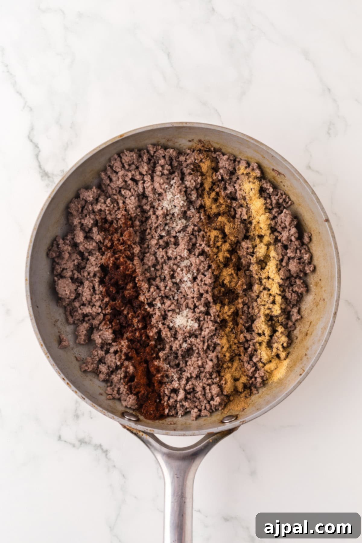 A skillet filled with cooked ground meat, arranged in lines are spices including chili powder, salt, cumin, and garlic powder, perfect for making a savory Cowboy Queso appetizer.