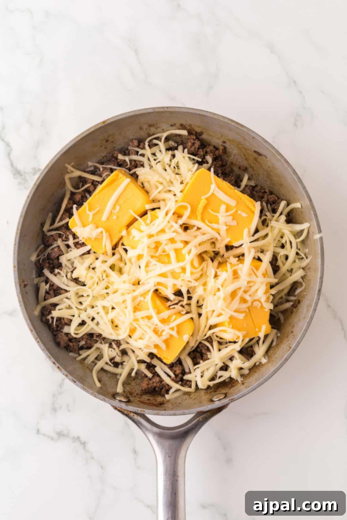 A skillet contains ground beef topped with shredded cheese and slices of processed cheese on a marble surface, perfect for your next Cowboy Queso dip.