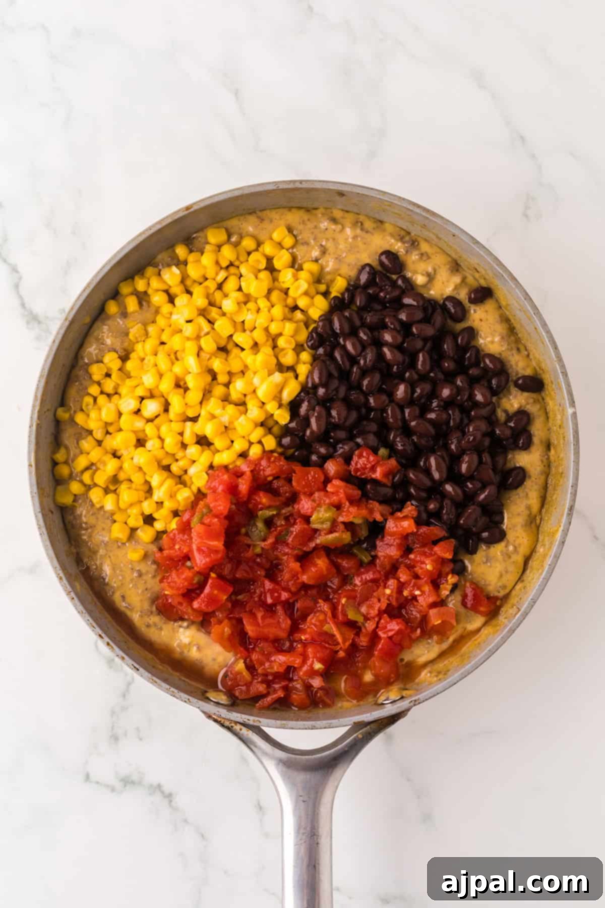 A skillet filled with mixed ingredients, including corn kernels, black beans, diced tomatoes, and a creamy base on a marble surface—truly a Tex-Mex delight.