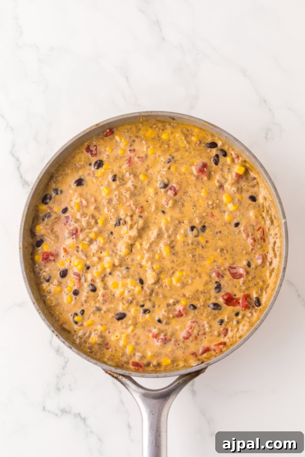 A skillet filled with a creamy mixture of corn, black beans, diced tomatoes, and seasonings, resembling a hearty Cowboy Queso dip, placed on a white marble countertop.