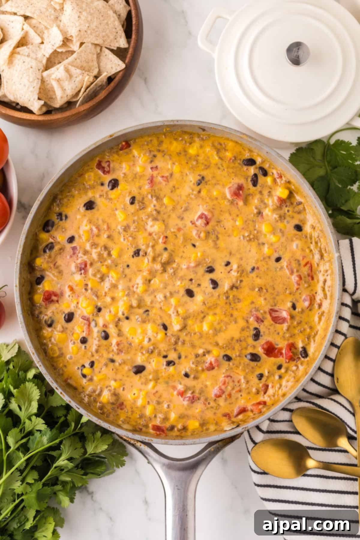 A skillet containing a creamy mixture of corn, black beans, diced tomatoes, and cheese—better known as Cowboy Queso. The surroundings include a bowl of tortilla chips, fresh cilantro, tomatoes, and a white pot lid.