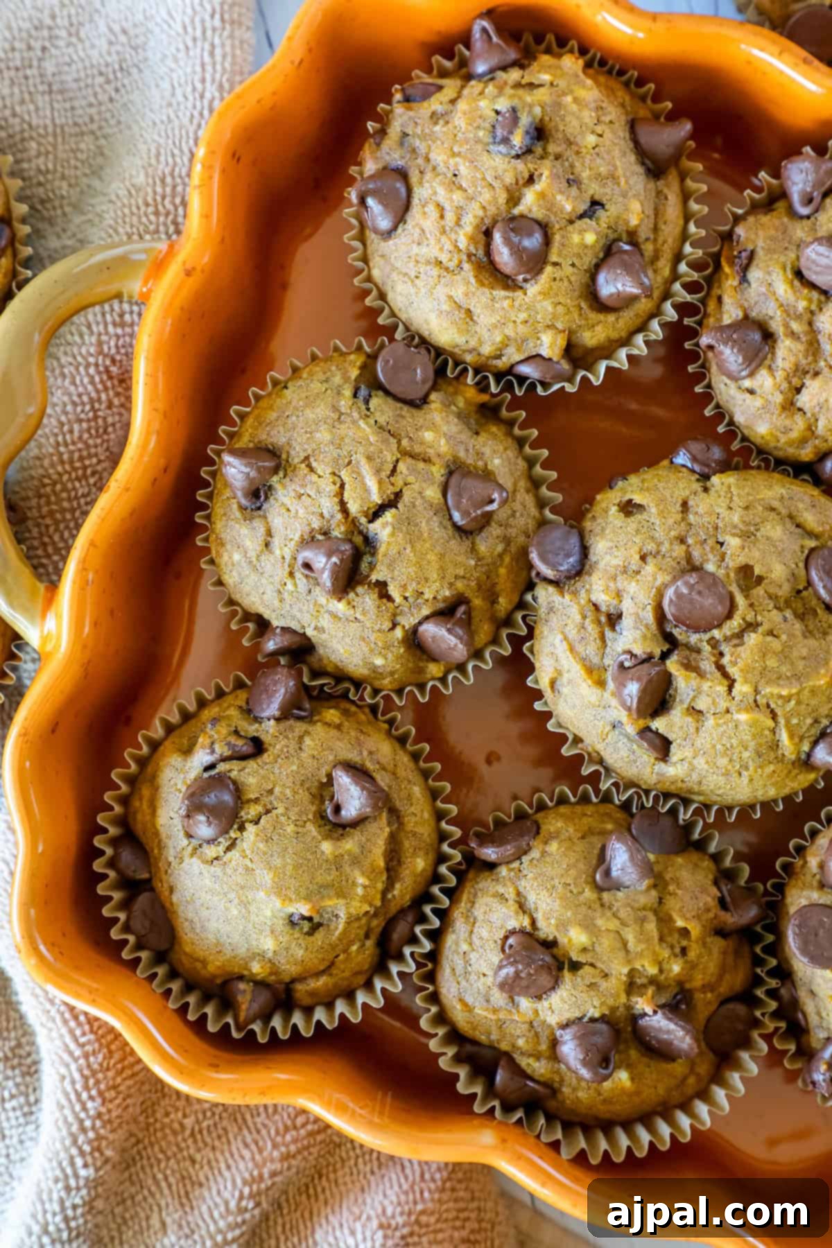 A ceramic dish holds six muffins topped with chocolate chips. The dish is orange with a handle, and it rests on a textured beige fabric.