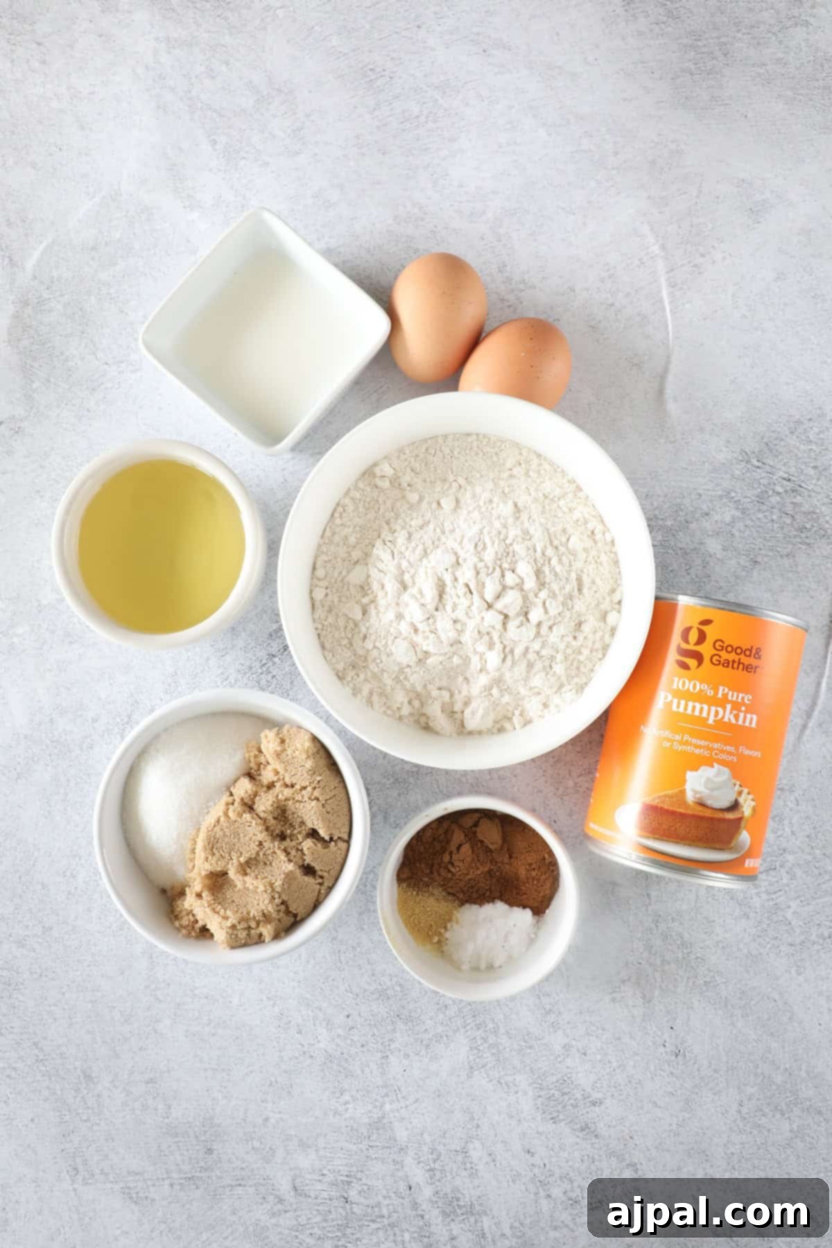Ingredients for baking on a grey surface: flour, eggs, milk, vegetable oil, canned pumpkin, brown sugar, white sugar, and spices in small bowls.