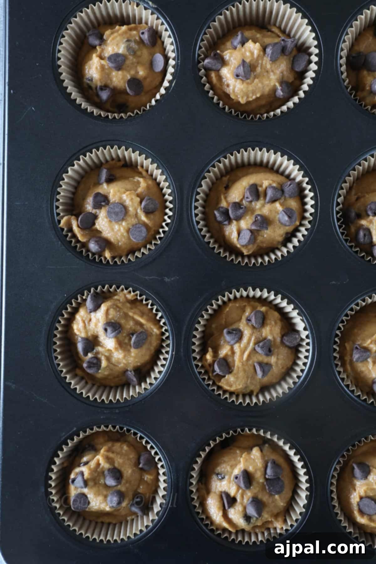A muffin tray with twelve paper liners filled with raw batter topped with chocolate chips, ready for baking.