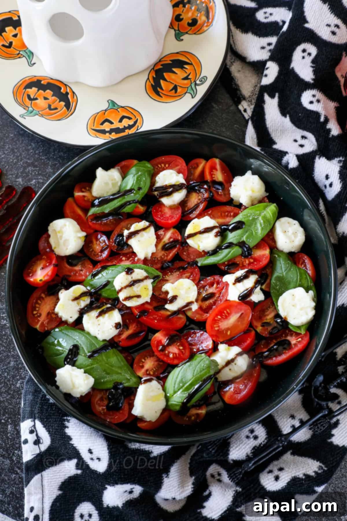 A bowl of Spooky Caprese Salad featuring halved cherry tomatoes, fresh basil leaves, and mozzarella shaped like pumpkins and ghosts. The background shows a Halloween-themed plate and a black cloth with ghost prints, setting a festive mood.