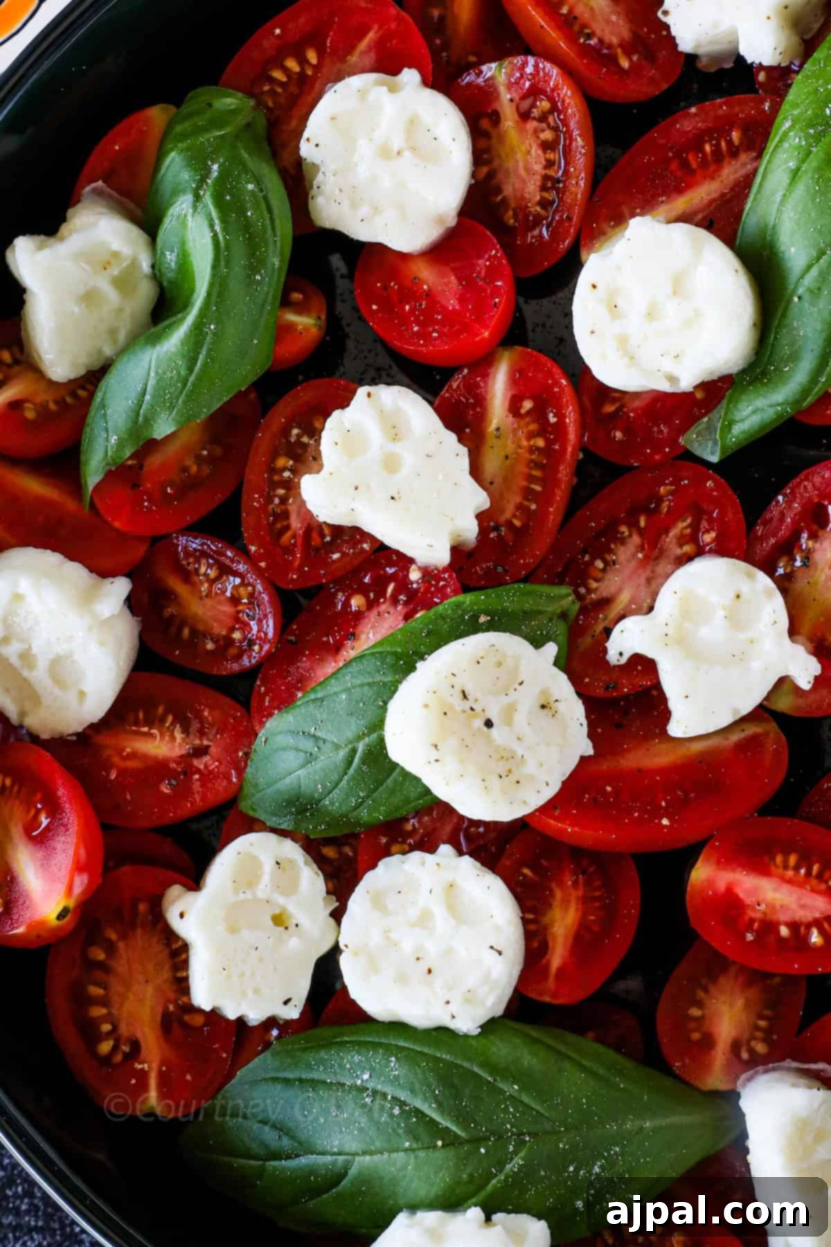 A close-up of a dish with halved cherry tomatoes, fresh basil leaves, dollops of mozzarella cheese, and a sprinkle of black pepper.