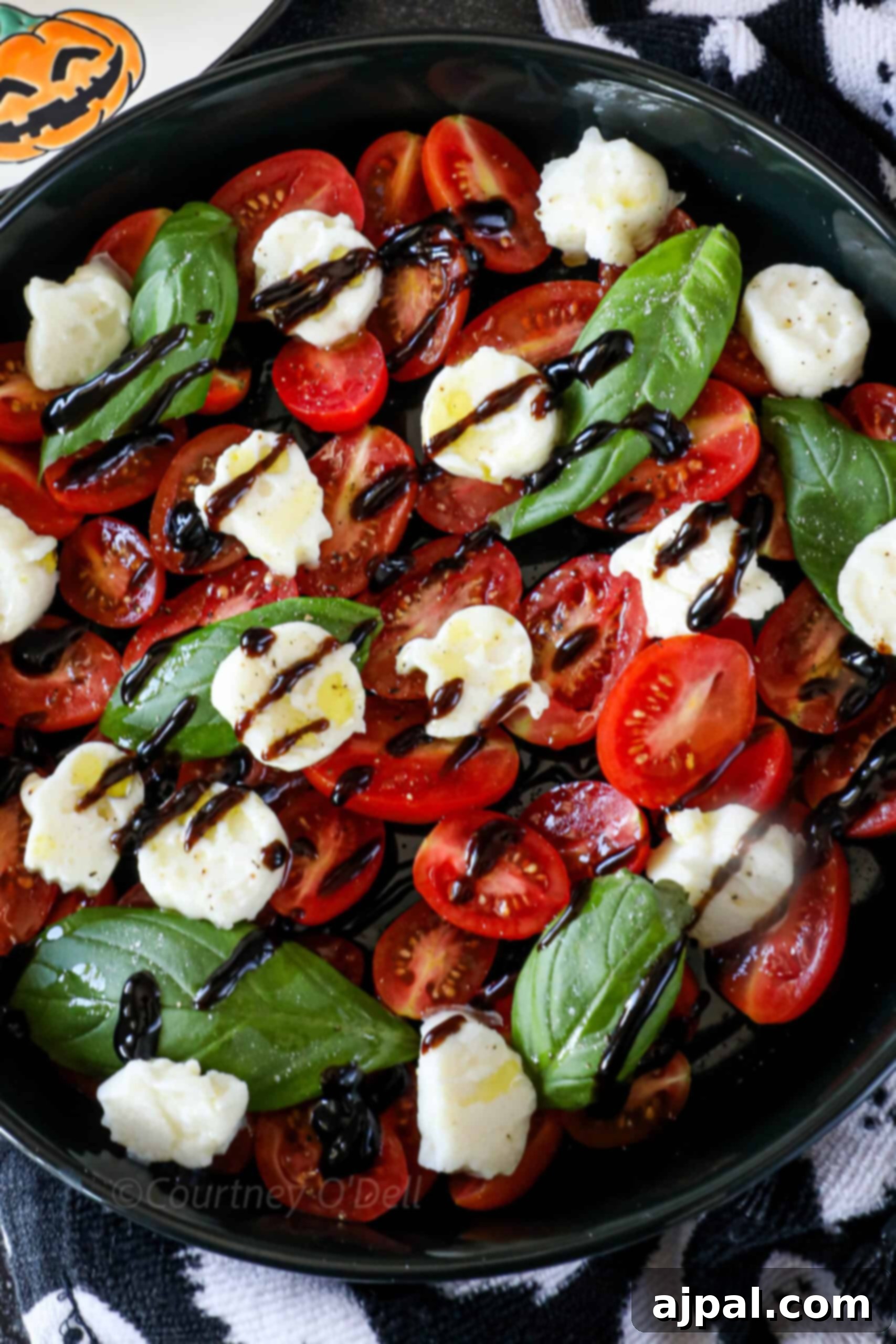 A vibrant Caprese salad featuring red cherry tomatoes, white mozzarella cheese shaped like ghosts, and green fresh basil leaves, all lightly drizzled with balsamic glaze. A Halloween-themed napkin adds a festive touch to the background.