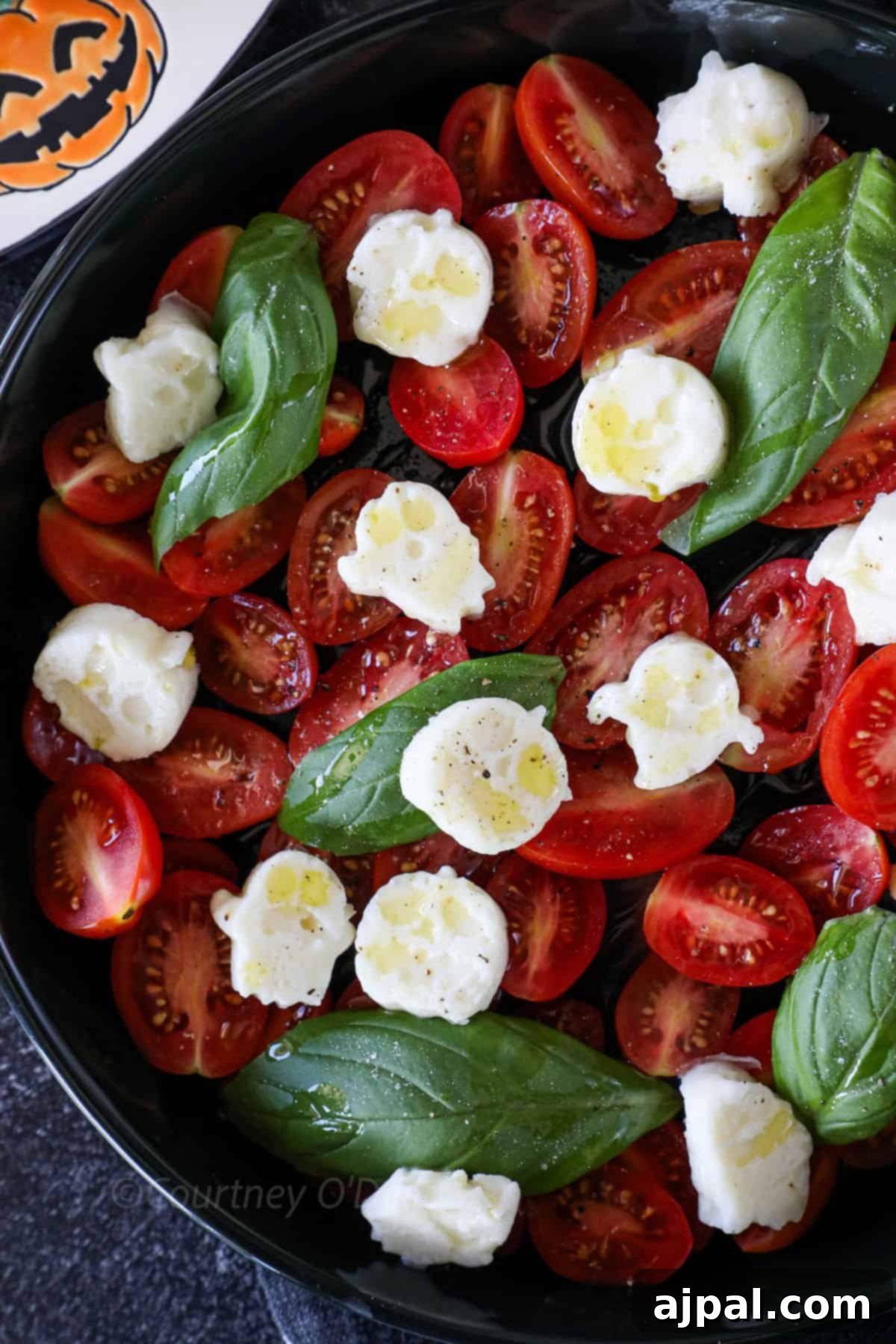 A bowl contains halved cherry tomatoes, fresh basil leaves, and dollops of mozzarella cheese, drizzled with olive oil. A Halloween-themed plate with a pumpkin face design is partly visible.