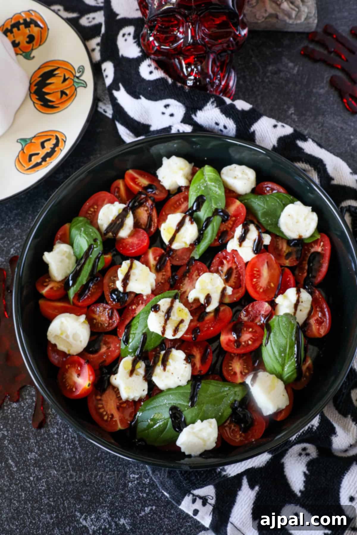 A beautifully presented bowl of Caprese salad, featuring vibrant cherry tomatoes, ghostly mozzarella shapes, and fresh basil leaves, all drizzled with balsamic glaze. In the background, Halloween-themed plates and napkins with pumpkin and ghost designs complete the festive setting.