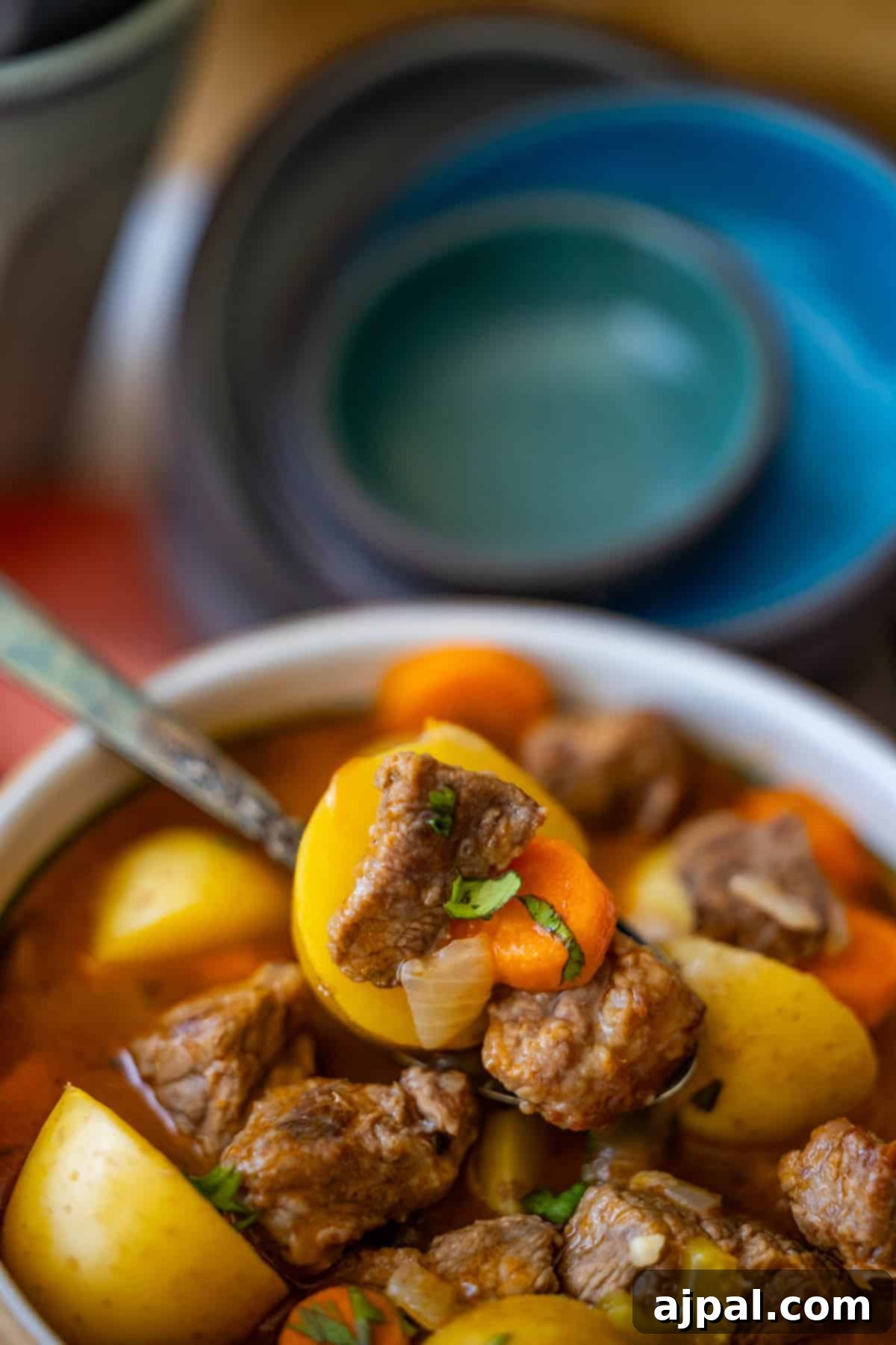 Simple Beef Stew 12 Close-up of a bowl of classic beef stew containing chunks of beef, potatoes, carrots, and onions, garnished with fresh herbs, with a spoon placed in the bowl. Stacked bowls are visible in the background.