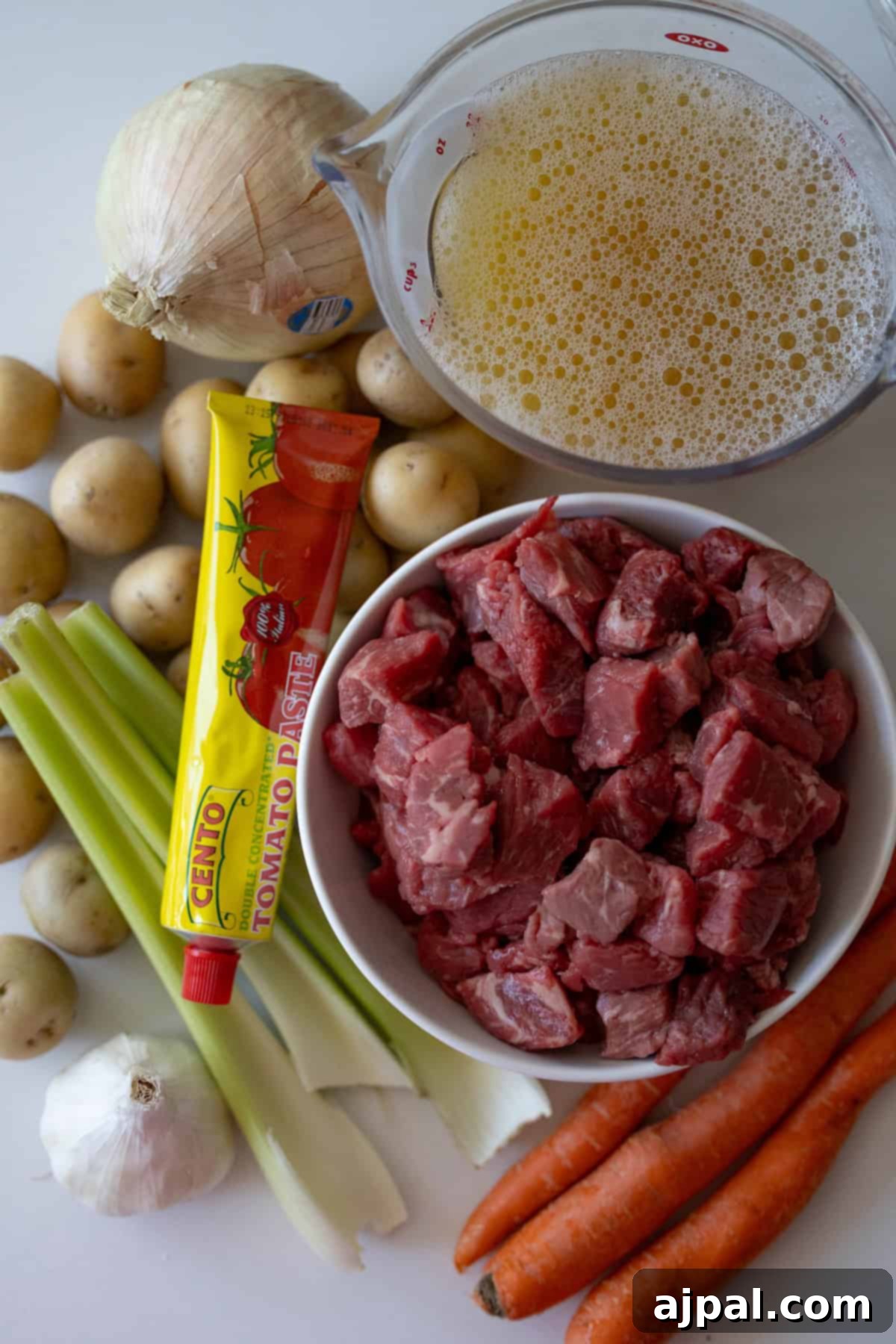 Simple Beef Stew 4 Ingredients for a classic beef stew include raw beef chunks in a white bowl, a tube of tomato paste, celery stalks, an onion, carrots, garlic, small potatoes, and a measuring cup filled with broth.