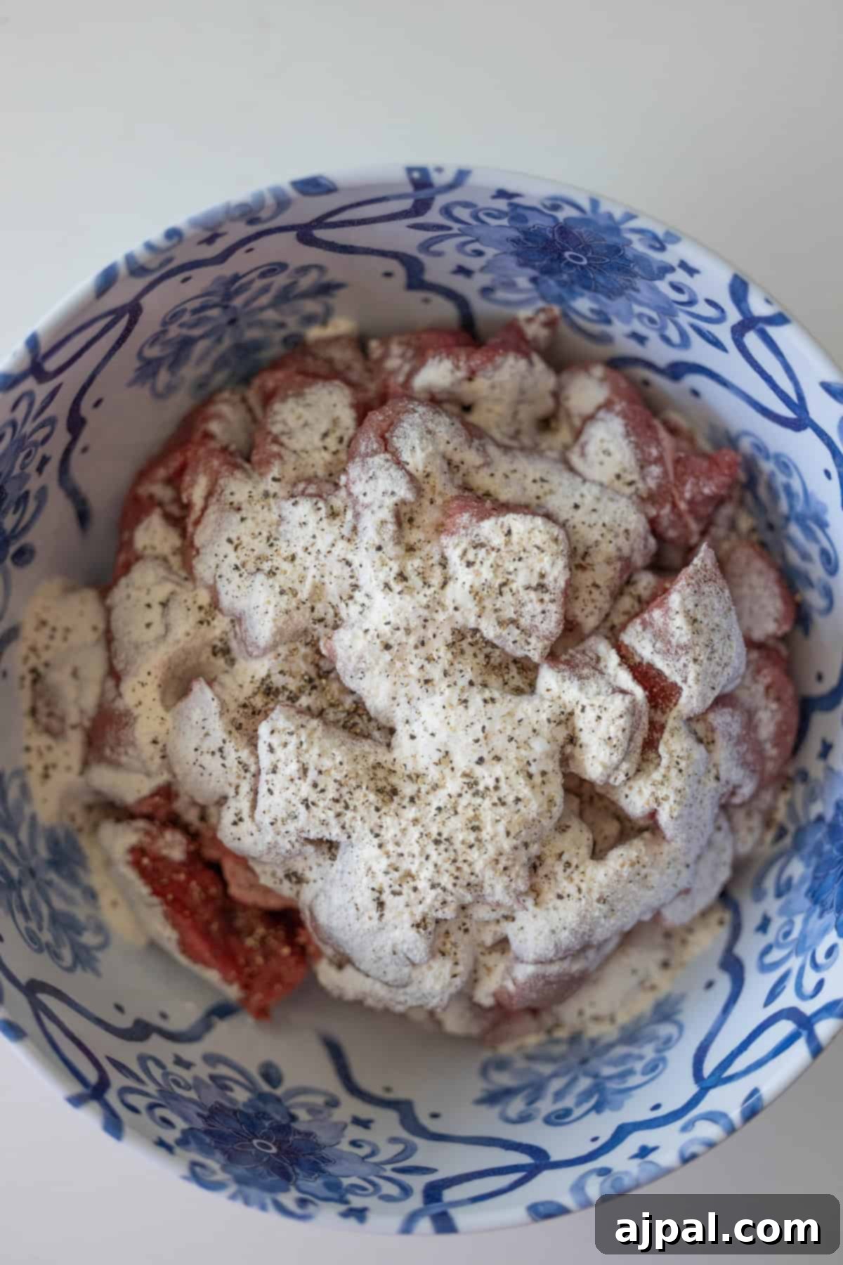 Simple Beef Stew 5 A bowl containing raw meat pieces covered with a white creamy marinade and sprinkled with black pepper suggests the beginning of a classic beef stew. The bowl, decorated with blue floral patterns, adds an elegant touch.