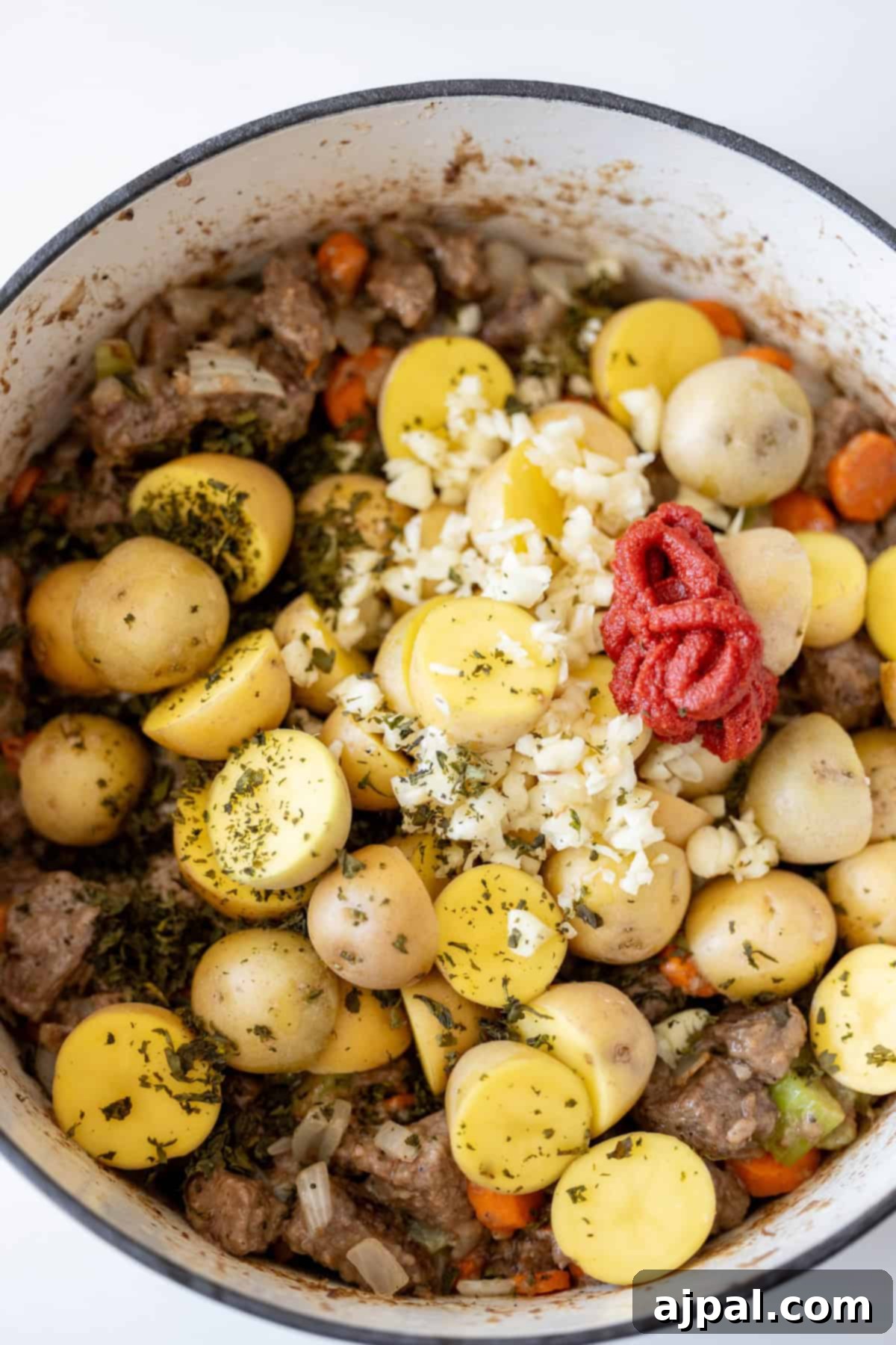 Simple Beef Stew 10 A pot contains yellow baby potatoes, minced garlic, tomato paste, diced carrots, and meat, all seasoned with green herbs for a classic beef stew.