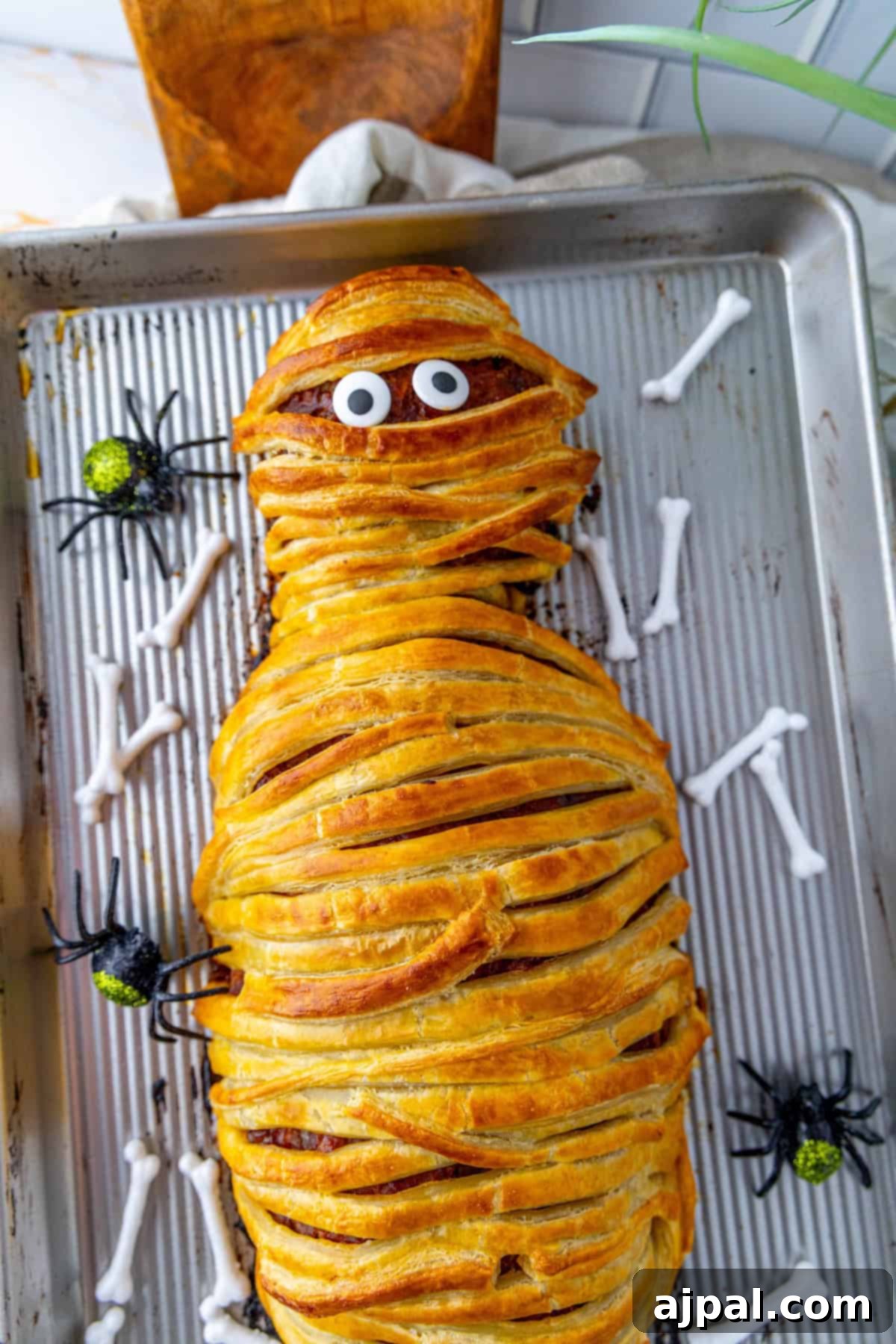 A festive mummy meatloaf wrapped in golden puff pastry, decorated with candy eyes, resting on a baking tray surrounded by plastic spiders and bone-shaped props, ready for Halloween.