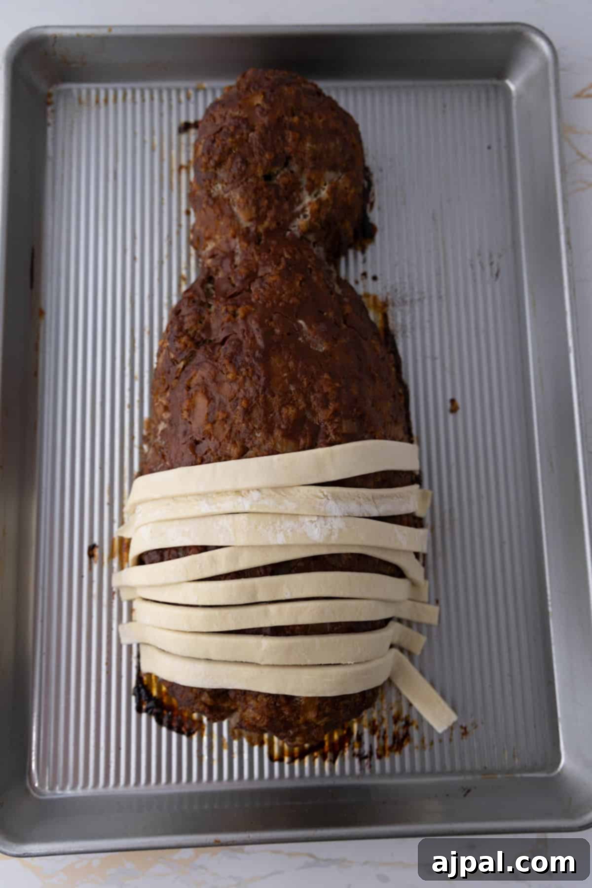 A mummy meatloaf shaped like a figure, partially wrapped in strips of dough on a baking sheet.