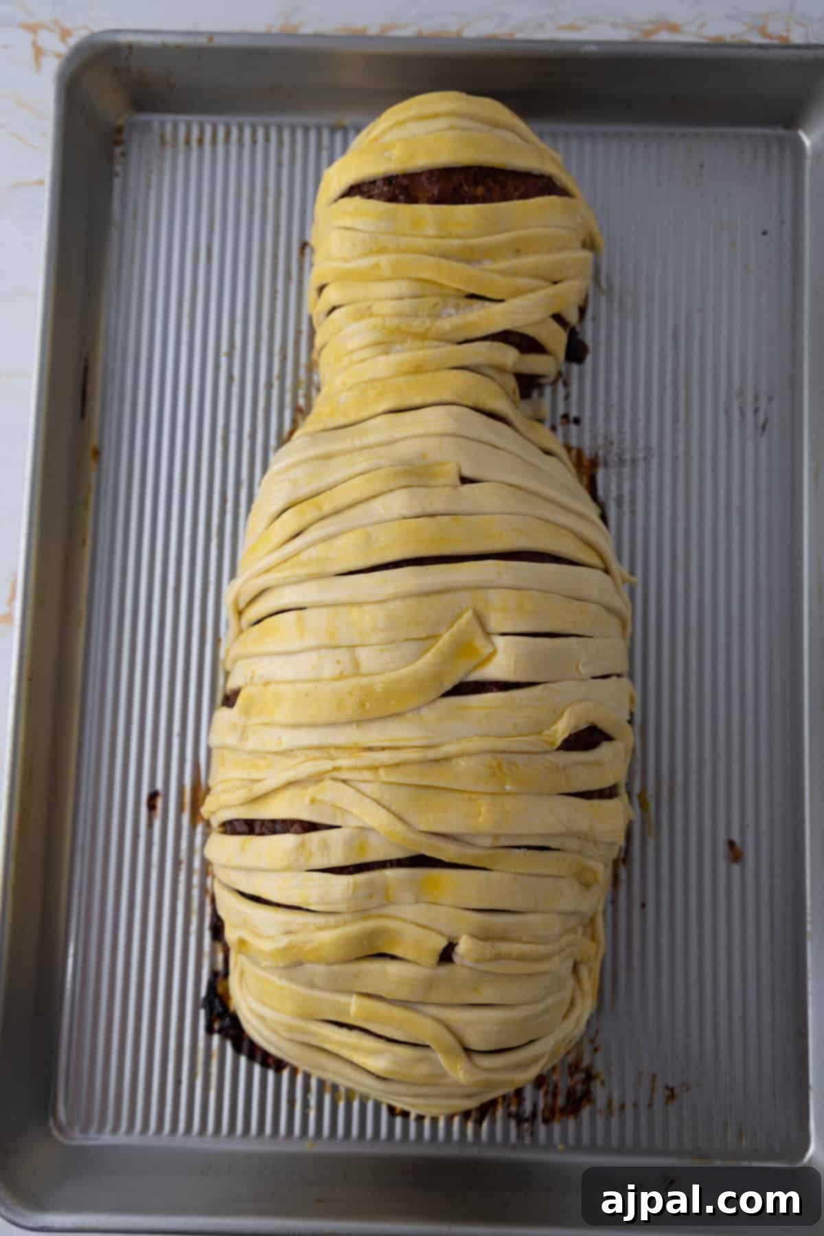 A mummy meatloaf fully wrapped in strips of puff pastry, positioned on a metal baking tray.
