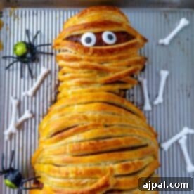 A mummy meatloaf dish wrapped in pastry with visible eyes, accompanied by plastic spiders and bones on a baking tray.