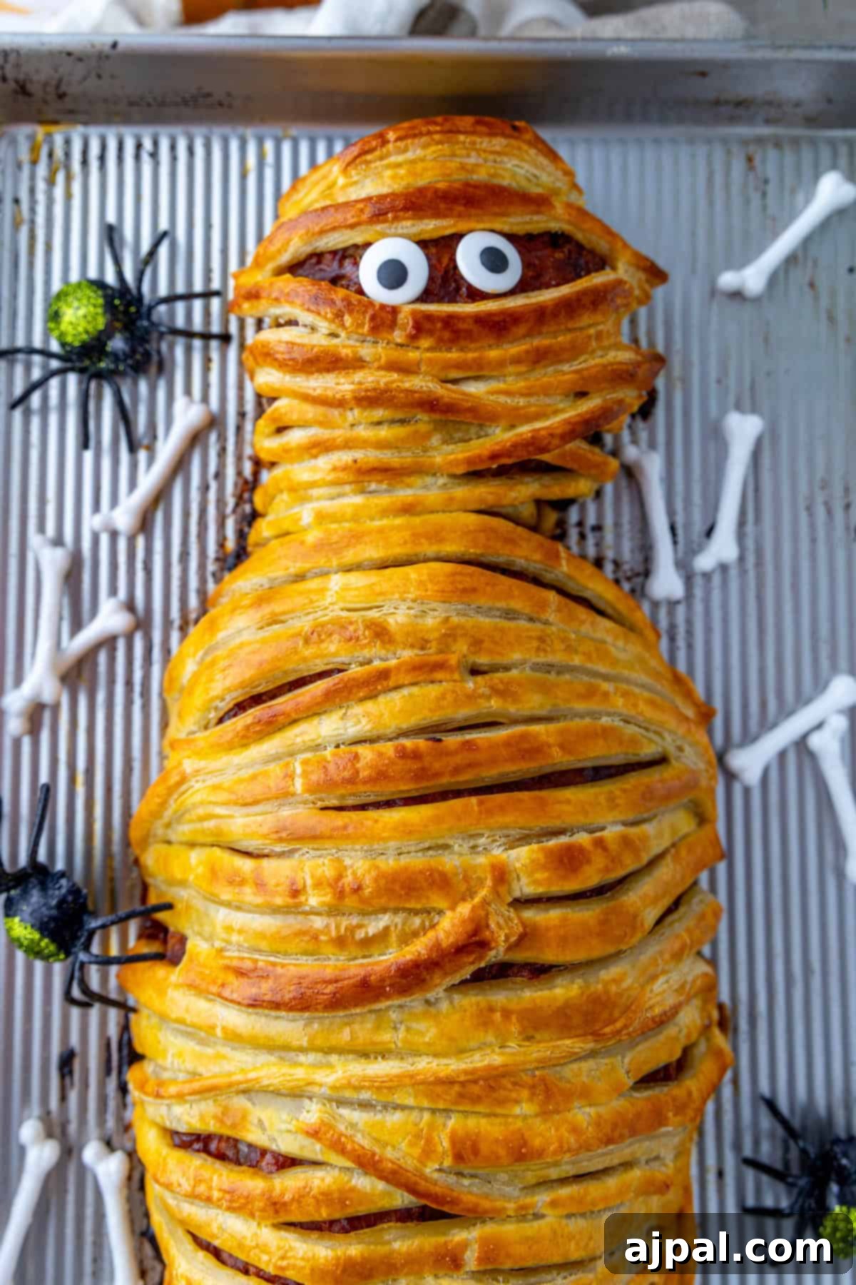 A perfectly baked mummy meatloaf, artfully wrapped in pastry 'bandages' with googly eyes, presented on a baking sheet amidst decorative plastic spiders and faux bone ornaments for a spooky Halloween display.