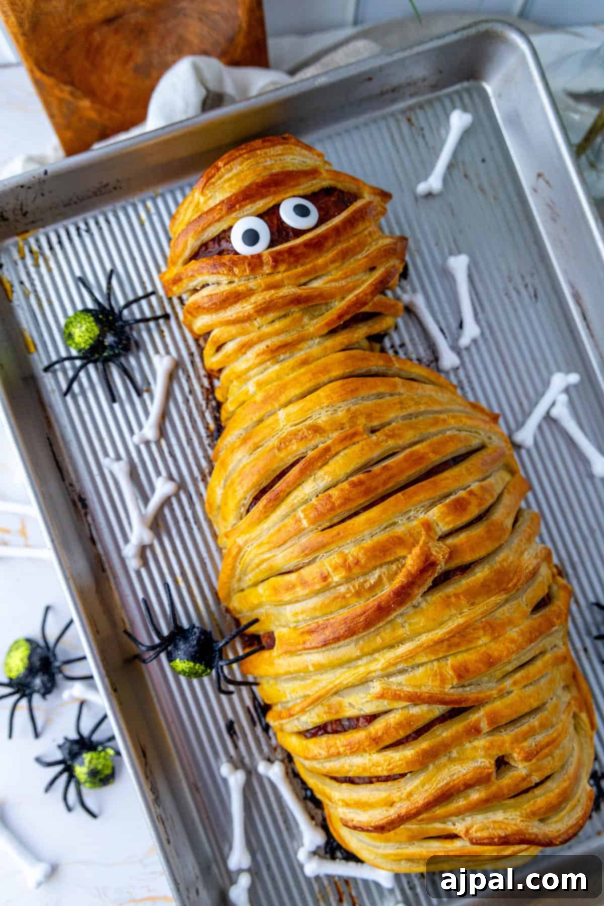 A playful mummy meatloaf with googly eyes on a baking tray, surrounded by decorative plastic spiders and bone-shaped treats, ready to be served for a fun Halloween meal.