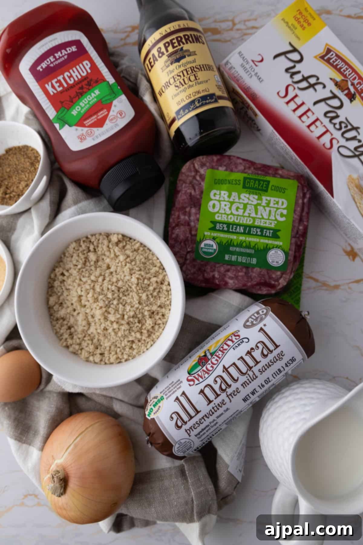 A variety of cooking ingredients, including ketchup, Worcestershire sauce, puff pastry, ground beef, sausage, onions, and milk, laid out for preparing a mummy meatloaf.