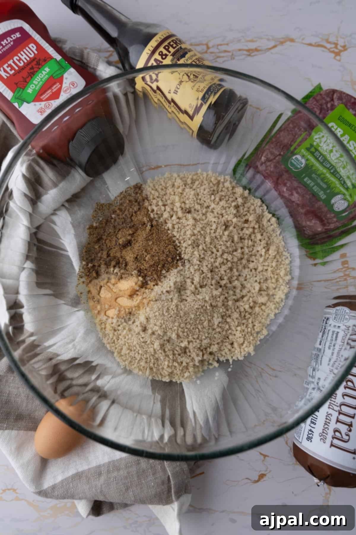 A glass bowl with cracker crumbs and spices, surrounded by ketchup, Worcestershire sauce, ground beef, egg, and sausage, all ingredients for mummy meatloaf.