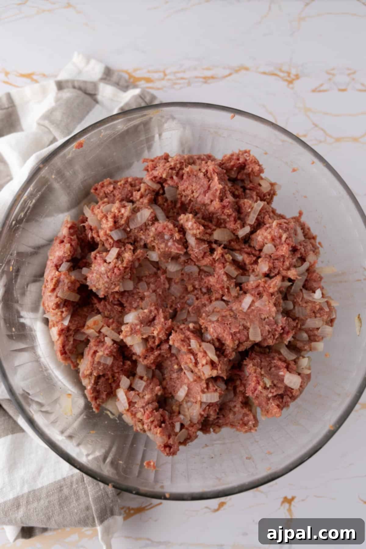 A glass bowl filled with a mixture of ground meat, diced onions, and seasonings for the mummy meatloaf.