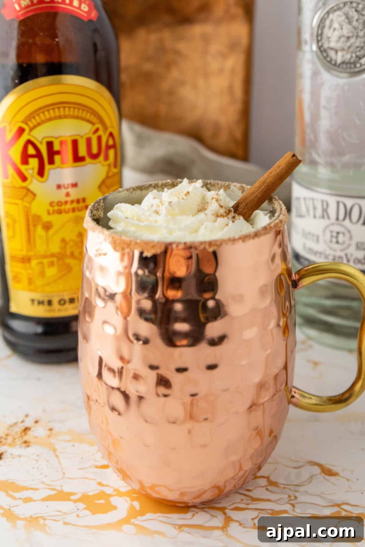Spiced Pumpkin White Russian 2 A copper mug filled with a whipped cream-topped beverage, garnished with a cinnamon stick, sits in front of a Kahlúa bottle and a bottle of Silver Dollar vodka.