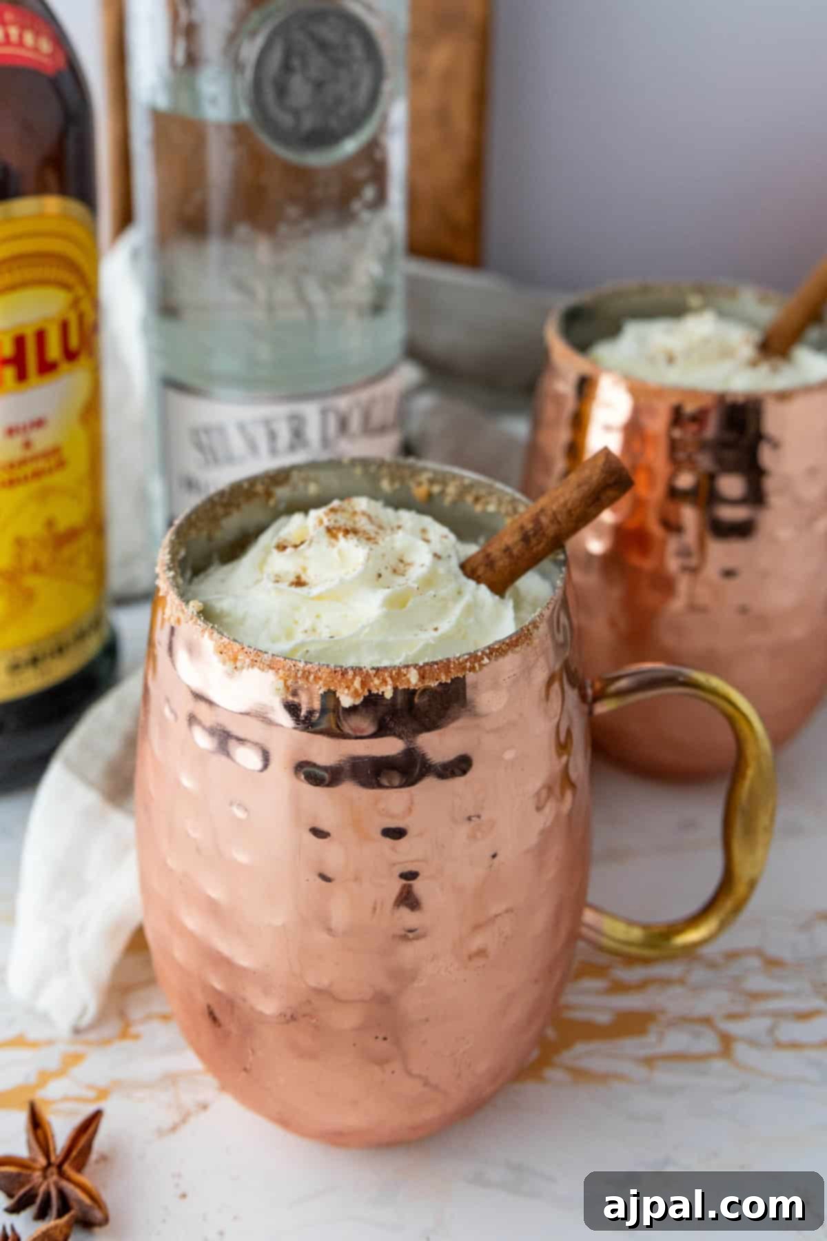 Spiced Pumpkin White Russian 3 Two copper mugs filled with creamy eggnog, topped with whipped cream and garnished with a cinnamon stick. Bottles of Kahlua and vodka are in the background on a light marble surface.