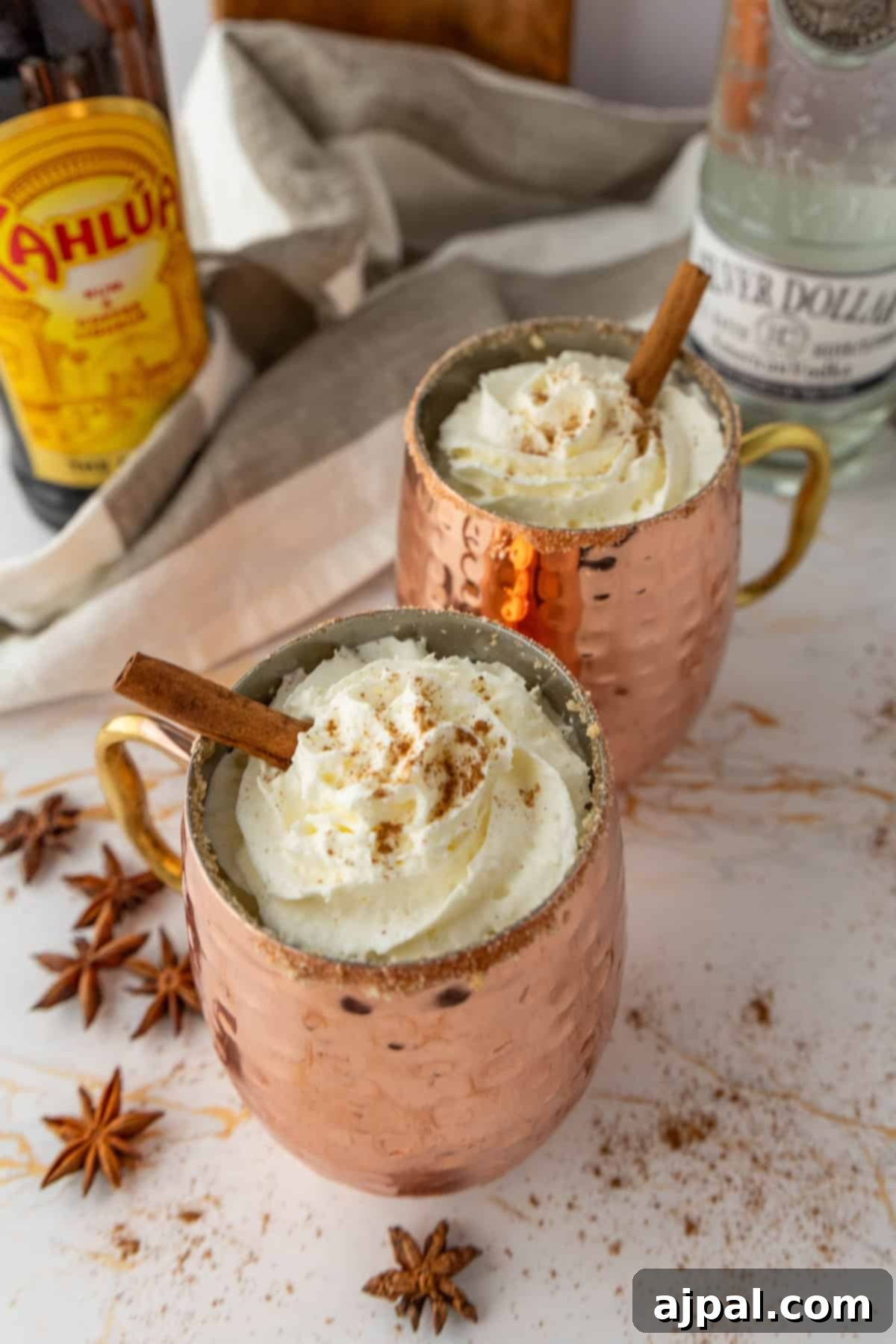 Spiced Pumpkin White Russian 4 Two copper mugs filled with a creamy drink topped with whipped cream and a cinnamon stick. A bottle of Kahlúa, a bottle of vodka, and star anise are in the background.