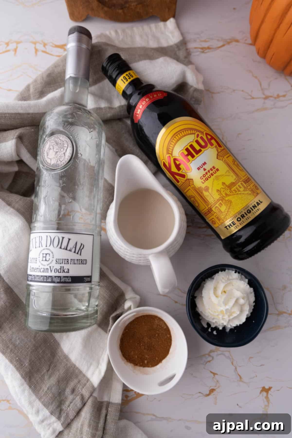 Spiced Pumpkin White Russian 5 Ingredients for a white Russian cocktail displayed on a marble surface: a bottle of vodka, a bottle of Kahlúa, a small pitcher of cream, a dish of whipped cream, and a bowl of cocoa powder.