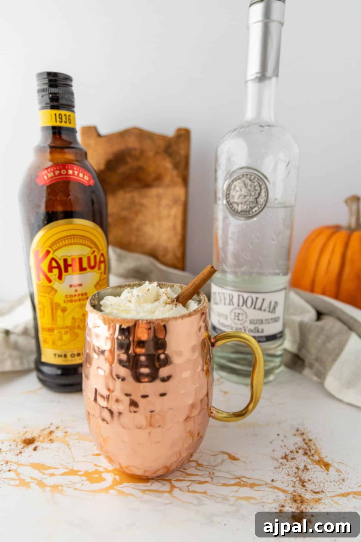 Spiced Pumpkin White Russian 6 A copper mug filled with a creamy drink, topped with whipped cream and a cinnamon stick is surrounded by a bottle of Kahlua, a bottle of vodka, and a pumpkin in the background.