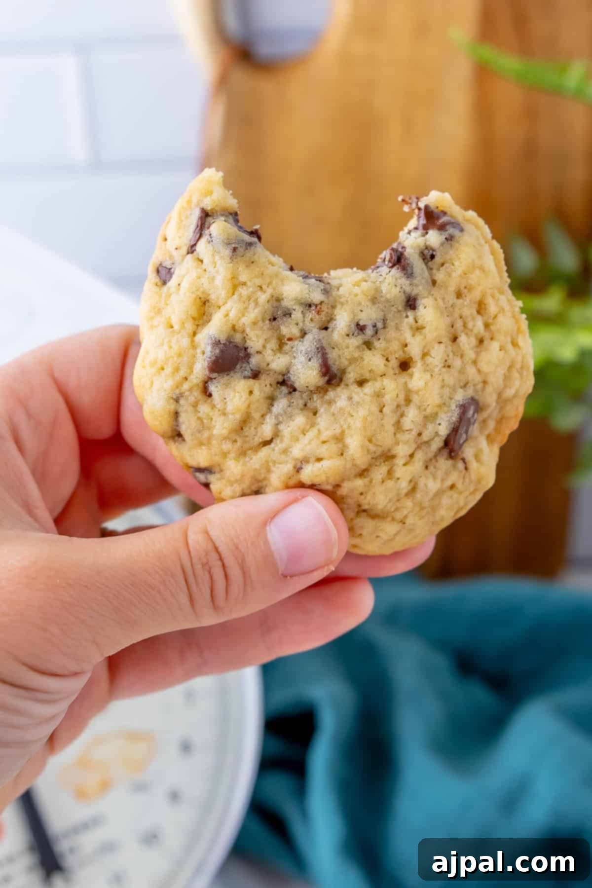 Next-Level Sourdough Discard Chocolate Chip Cookies 12 A hand holds a partially eaten sourdough chocolate chip cookie with a blue cloth and cutting board in the background.