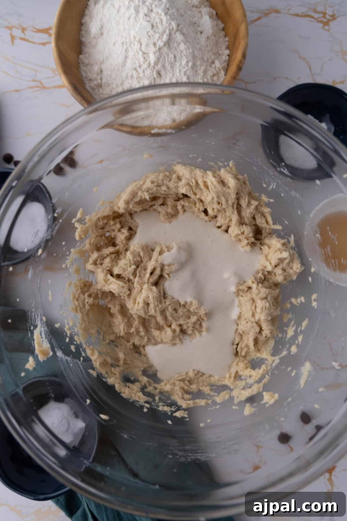 Next-Level Sourdough Discard Chocolate Chip Cookies 7 A glass bowl contains partially mixed cookie dough with a liquid mixture being added. In the background, there is a wooden bowl filled with flour, hinting at the delicious potential of sourdough chocolate chip cookies.