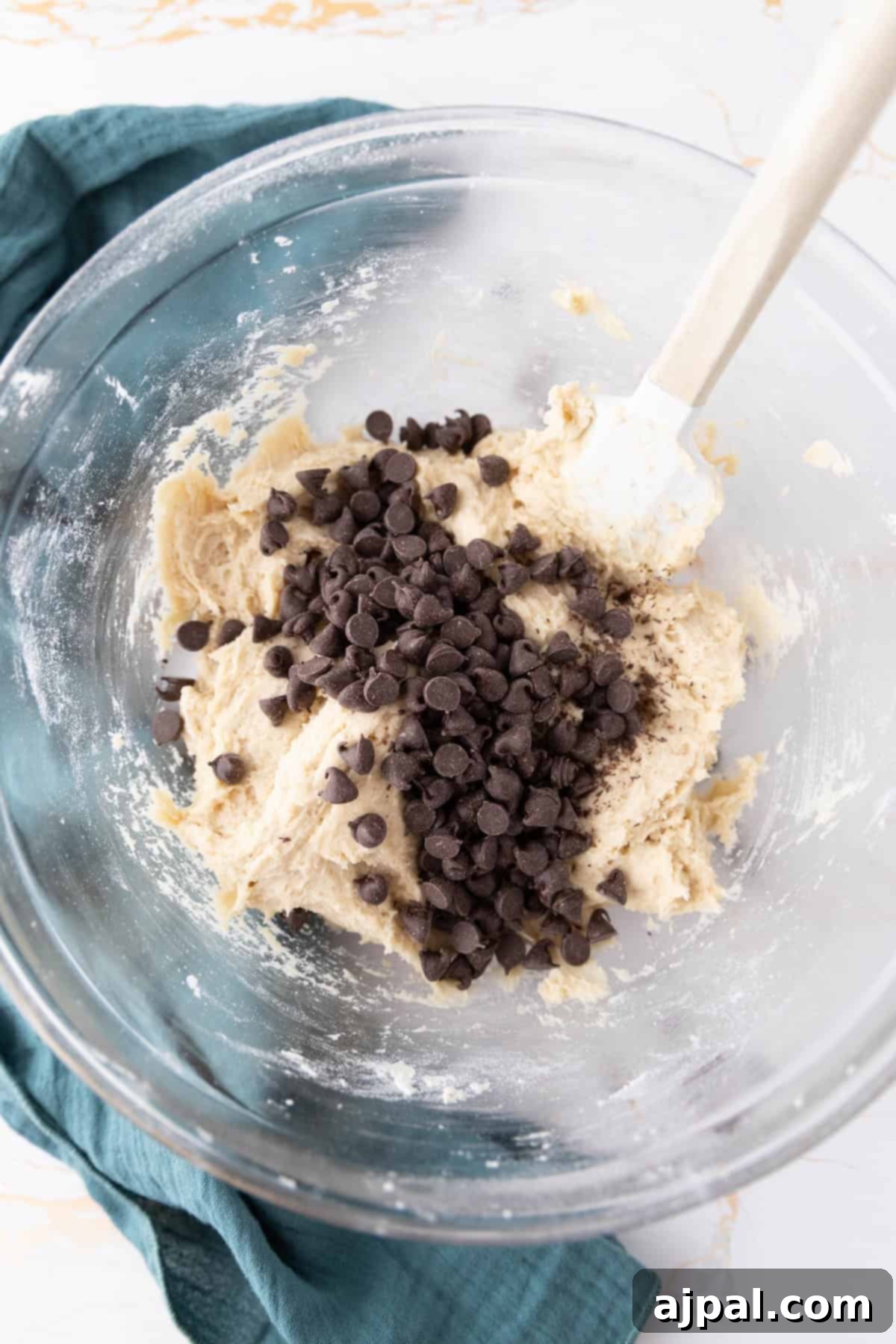 Next-Level Sourdough Discard Chocolate Chip Cookies 10 A glass bowl containing sourdough chocolate chip cookie dough with chocolate chips on top, and a white spatula is resting in the mixture. A teal cloth is partially visible under the bowl on a marble surface.