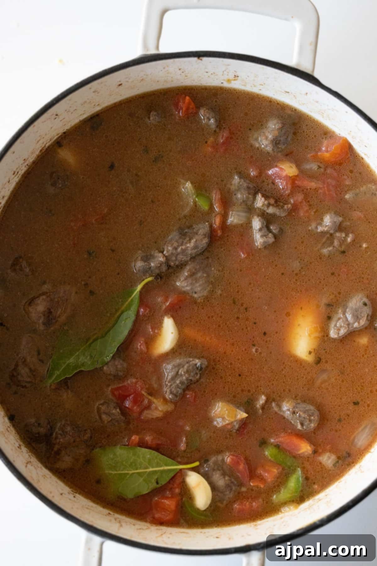 A pot of beef stew featuring generous chunks of meat, sliced tomatoes, whole garlic cloves, a bay leaf, and visible spices simmering in a savory broth.